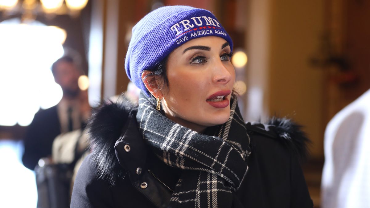 Laura Loomer, a white woman, wears a blue knit hat with white words that read "Trump - Saves America Again." She is also wearing a black parka and a black and white tartan scarf.