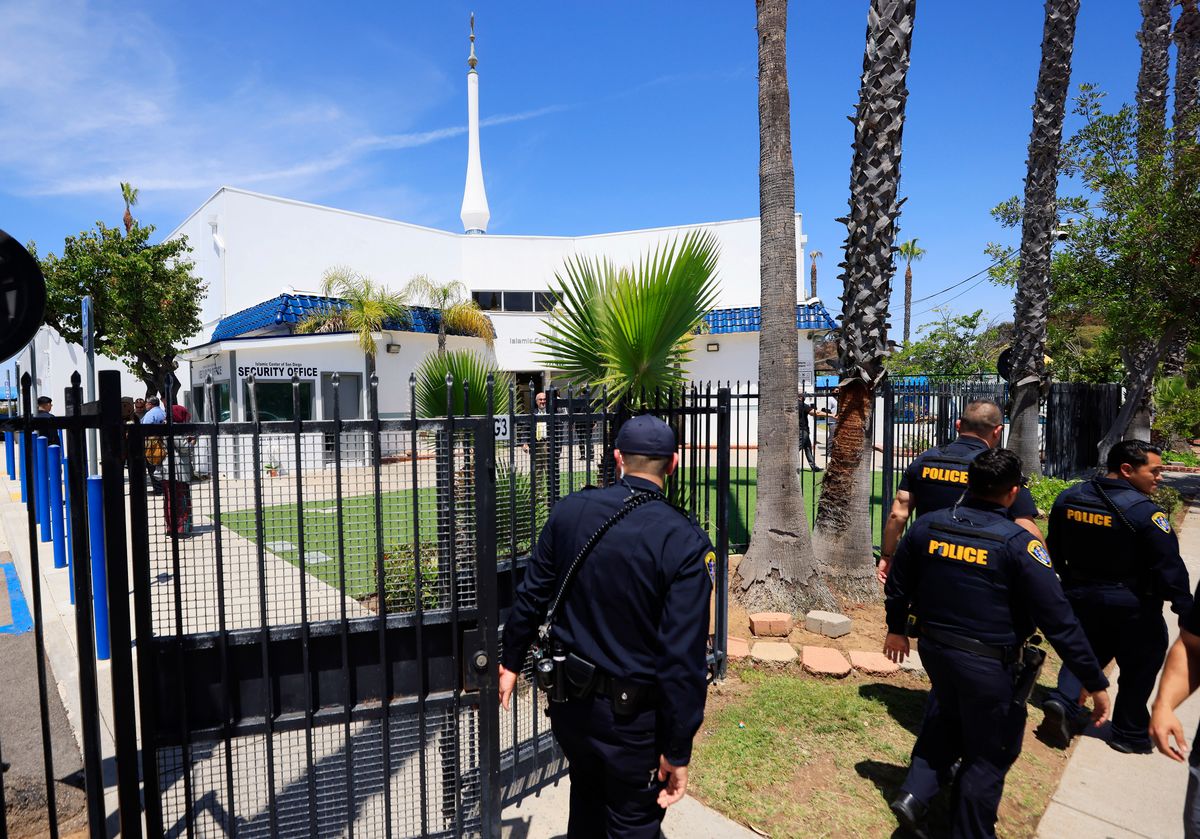 People wearing blue and yellow police uniforms walk in front of a gated white mosque.