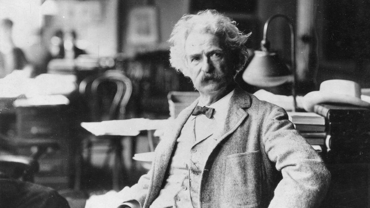 A black and white photo shows American author Mark Twain posing for a picture in a chair while wearing a bow tie and coat, with other out of focus furniture visible in the background.