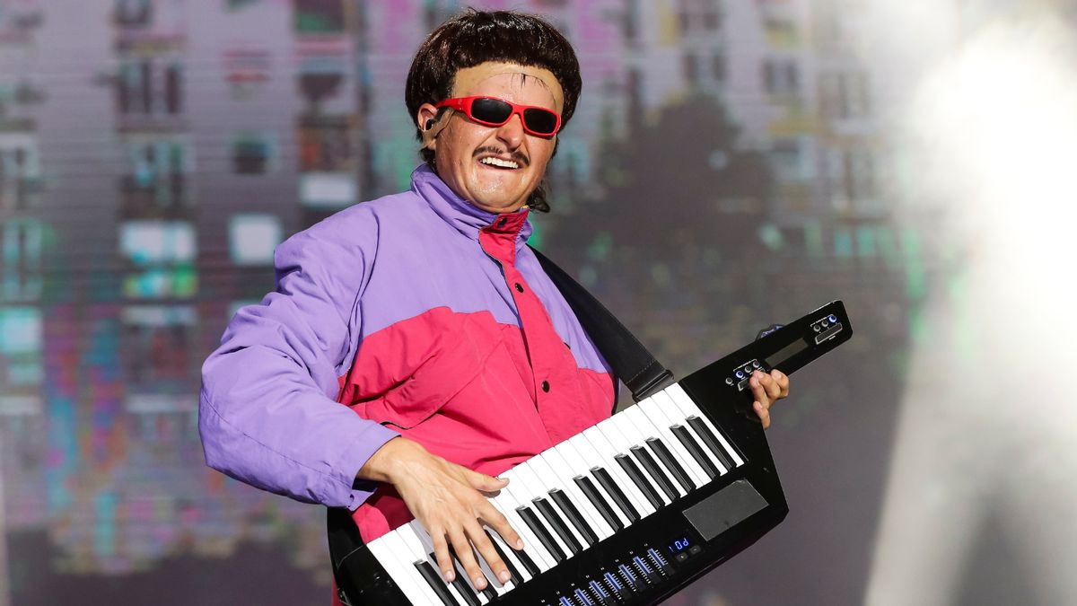 Oliver Tree, a young white man, wears a black, bowl-cut wig, goofy red sunglasses and a neon pink and purple puffer jacket while playing a keytar.