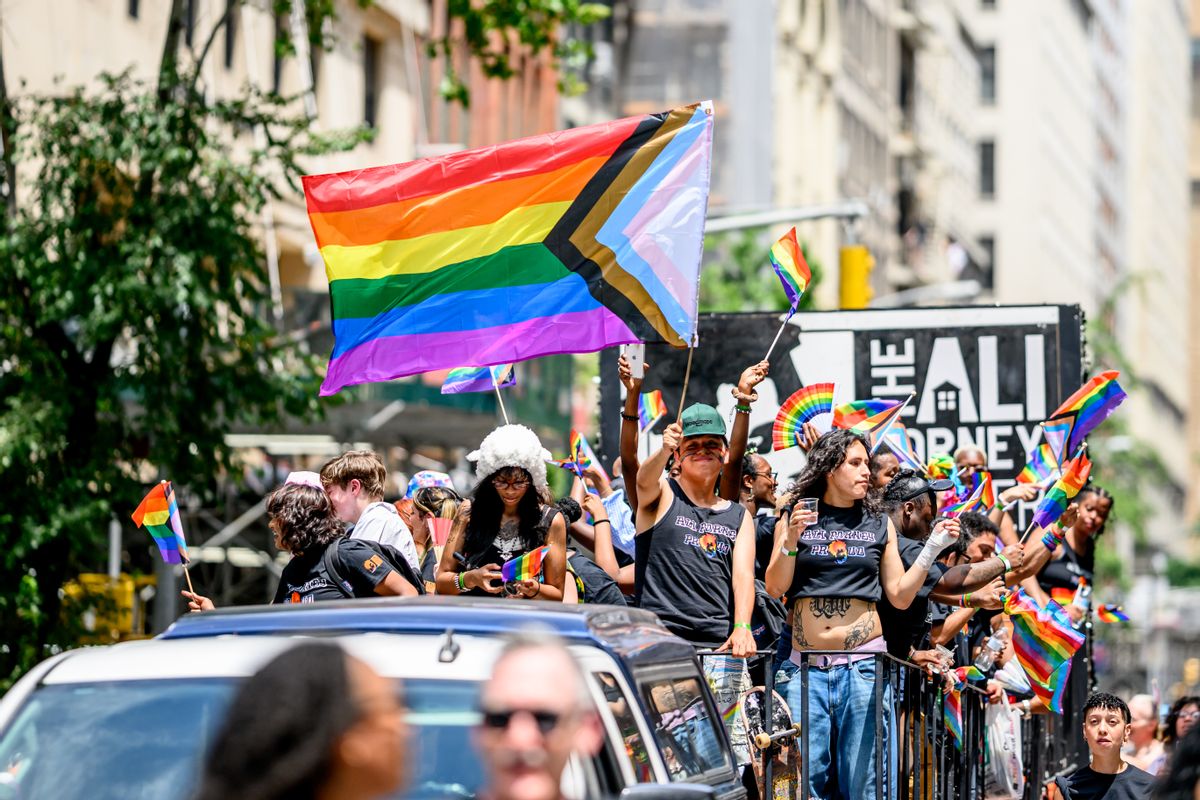 A person holding a large rainbow pride flag with white, pink, blue, brown and black chevron stripes stands among many other people holding smaller pride flags. 