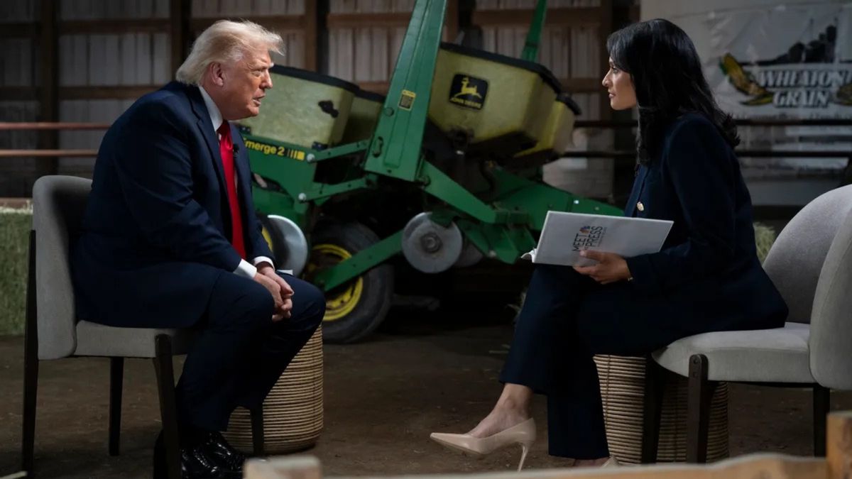 Trump leans forward in his seat while speaking to Kristen Welker. They appear to be sitting in a barn of sorts and a large piece of John Deere farm machinery is visible behind them. 