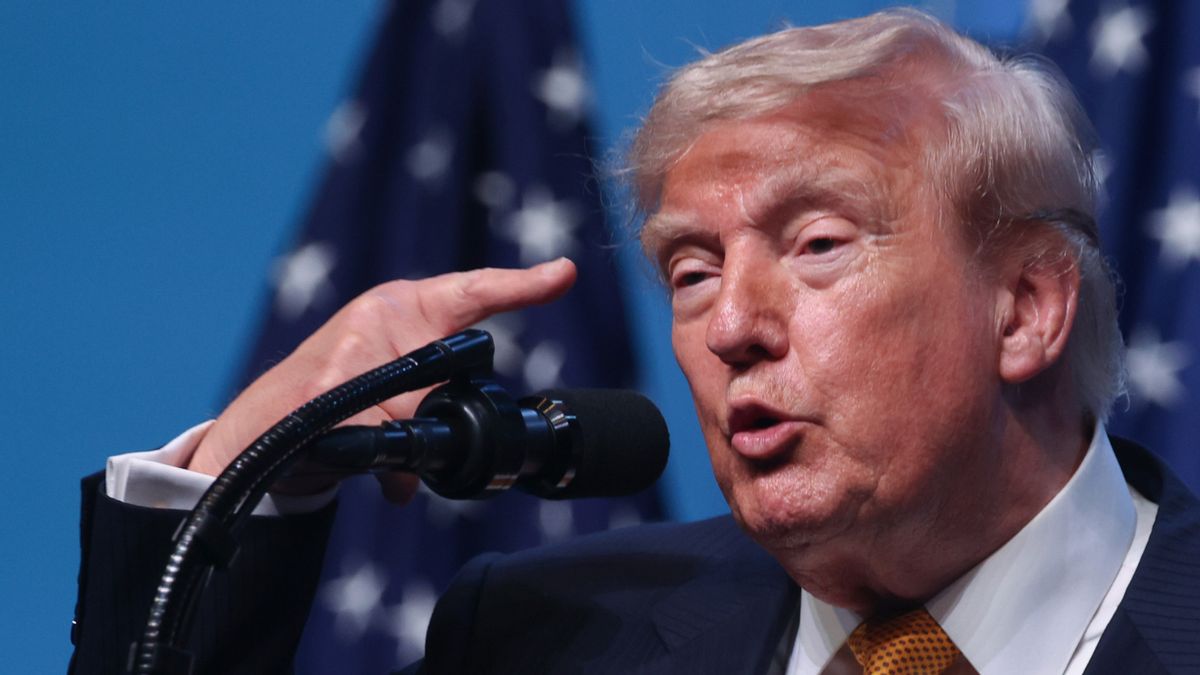 U.S. President Donald Trump raises a finger to his head while speaking in front of a microphone. 
