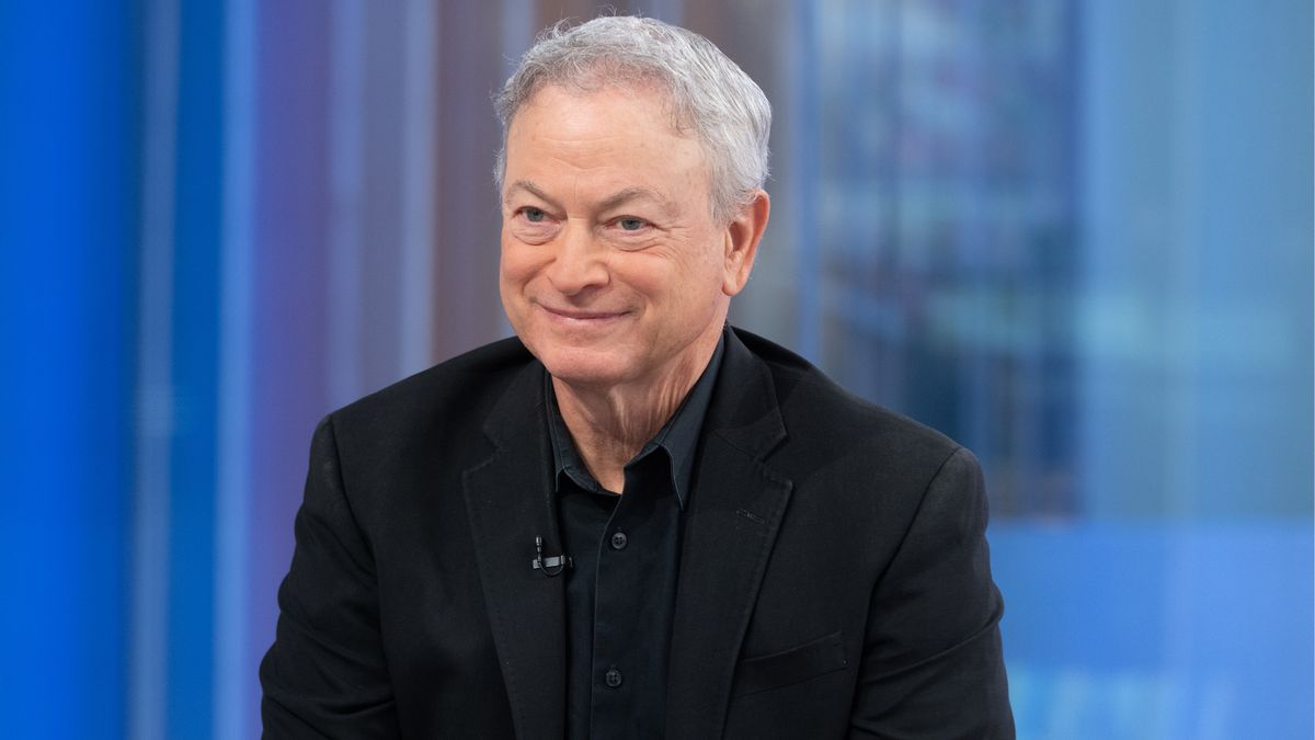 Actor Gary Sinise, a white man with gray hair, wears a black suit and smiles while sitting in front of a blue background.