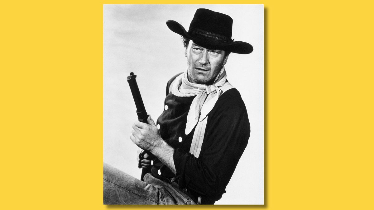 Black-and-white image shows actor John Wayne seated in a cowboy hat while holding up a gun. 