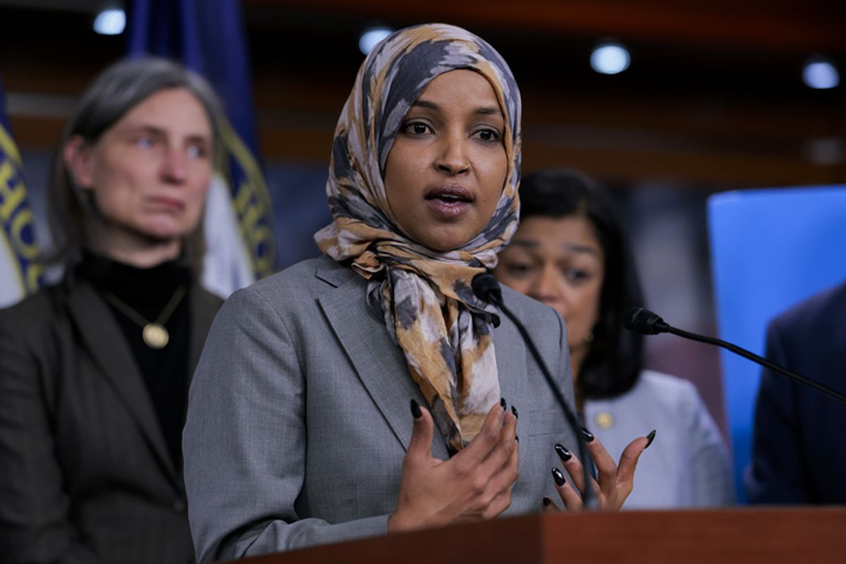 U.S. Rep. Ilhan Omar, a Somali woman in a gray suit and gray and yellow headscarf, speaks at a podium. 