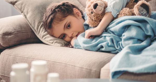 Child laying on couch covered by blanket hugging teddy bear with pill bottles in the foreground