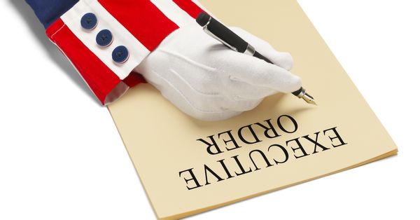 Hand in white glove and red, white, and blue cuff signing a piece of paper that says: EXECUTIVE ORDER.