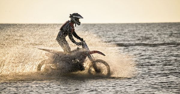 A rider on a cross-country motorcycle rides by the sea and raises a spray of water.