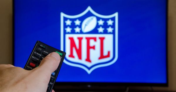 Hand holding remote in front of television screen displaying the NFL logo.