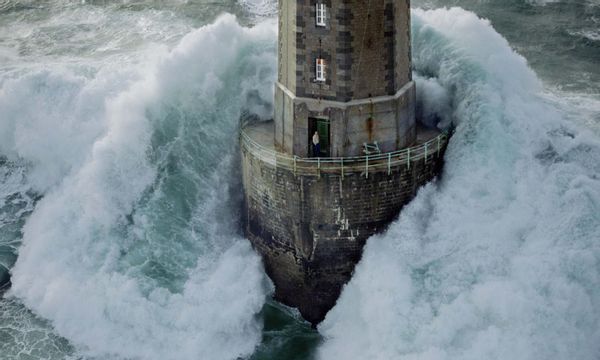 A photograph shows a big wave hitting a lighthouse while a lighthouse keeper stands in the doorway and the picture is from 1989 at the La Jument Lighthouse in France.