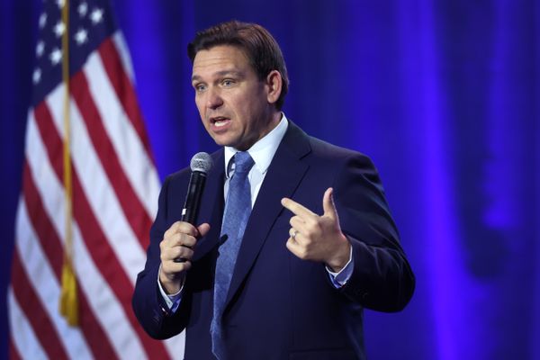 A white man speaking in a microphone stands near a blue backdrop with an American flag next to it.