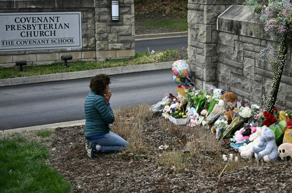 A person kneals in front of front of flowers and stuffed animals left at a gate. Next to the gate reads a plaque that says, "Covenant Presbyterian Church - the Covenant School."