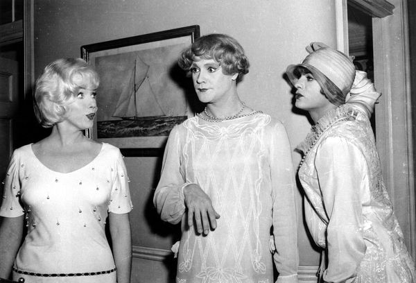 Three white people stand near a wall. The two on the right are wearing dresses. 