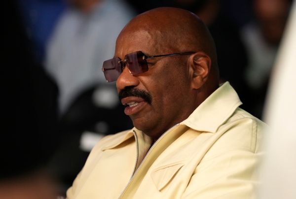 A Black man wearing sunglasses and a yellow shirt is sitting down. 