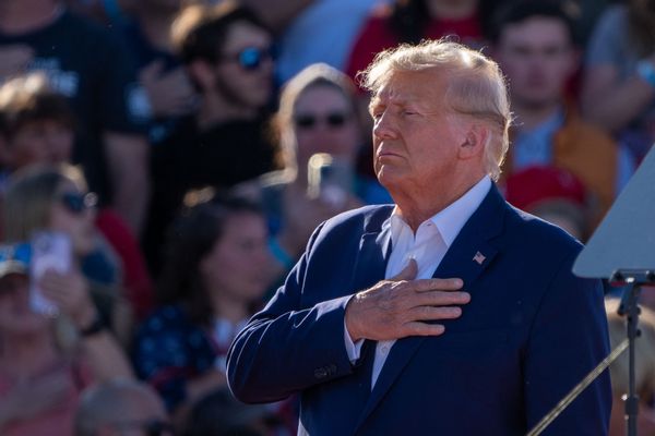 A white man wearing a blue suit with an American flag pinned on it stands with his hand on his hear. Many people are photographed behind him.