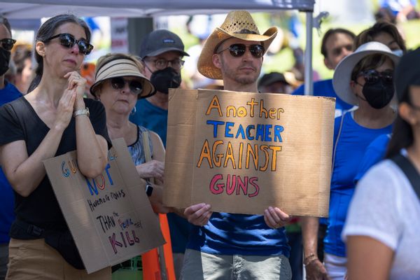 People stand gathered in a crowd. One wearing a cowboy hat says, "Another Teacher Against Guns."