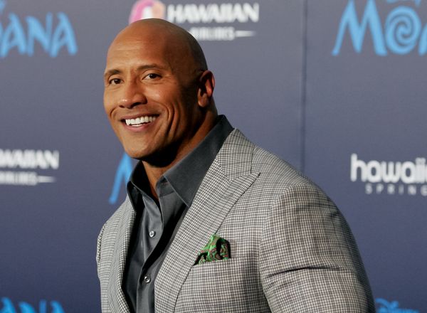 A Black/Samoan man in a gray suit smiles looking towards the camera.