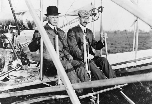 Two white men sit at the controls of an airplane in a black and white photograph.