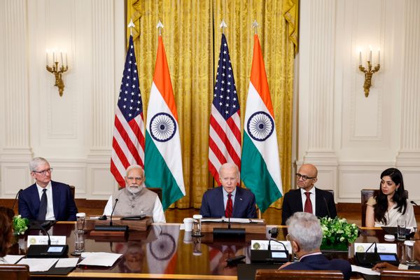 A white man sits in the middle of a table, surrounded by people on either side of him. In the background are two flags with red and white stripes and white stripes on a blue square, as well as two flags with orange, white and green stripes. On the white stripe of the flag is a spoked wheel.