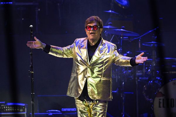 A white man wearing a gold suit and black glasses with orange lenses has his hands outstrethed while standing on a stage.