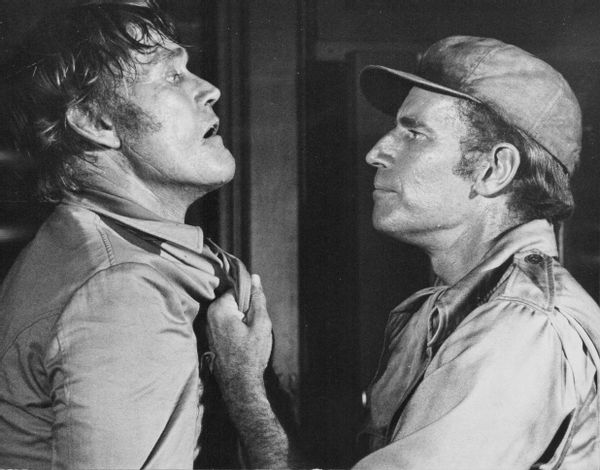 A white man holds another white man by the collar of his shirt.