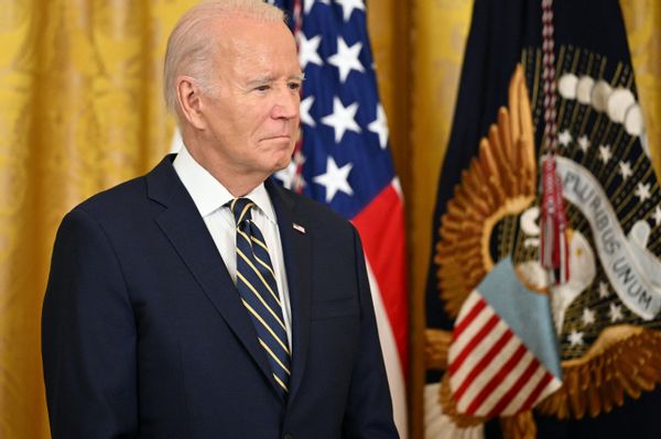 A white man in a blue suit stands next to two flags against a yellow curtain.