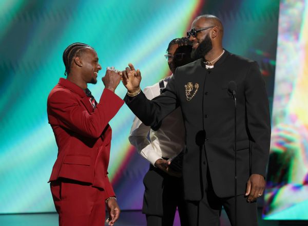 Two Black men smile at each other while linking pinkies. The one on the left is wearing a red suit, while the one on the right is wearing a black suit. A Black boy can be seen faintly behind the Black man on the right.