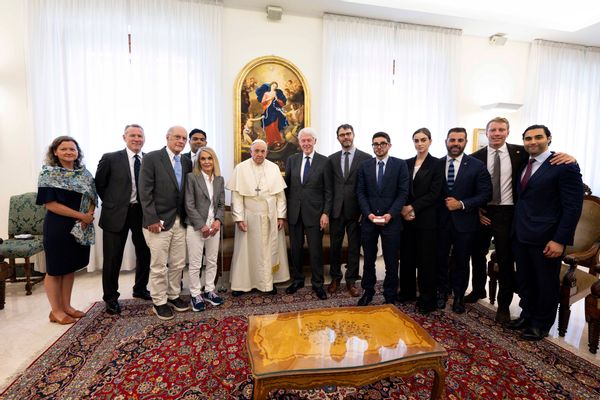 Several people gather together in a room to take a photo together. Many on the right are wearing black suits, while on the middle left a man wears all white with a cross around his neck.