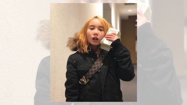 A white girl with blond hair wearing a black jacket and a stack of cash next to her right cheek is standing in a hallway.