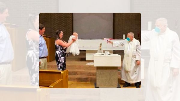 A priest wearing a mask points a water gun at a baby and their family.