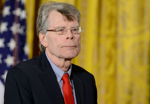 A white man wearing a suit and glasses is standing in front of a yellow backdrop. You can see the American flag behind him on his left side.