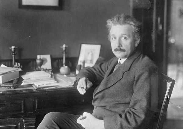 A black and white photo shows a white man sitting at a desk. His hair is white and gray, and he has a moustache. The desk is full of papers.