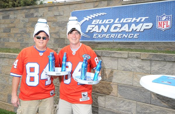 Two white people wear orange jerseys in front of a sign that says "Bud Light Fan Camp Experience," with the NFL logo next to them. One of the people is holding Bud Light bears on platters.
