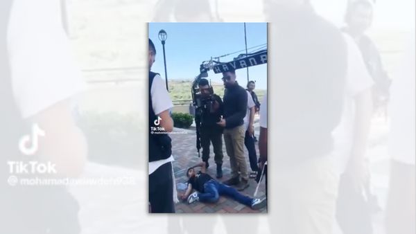 A Palestinian boy lies on the ground. People stand around him . You can see a film camera above him.