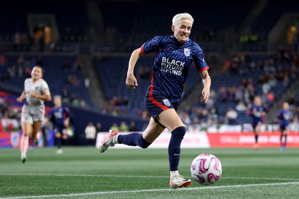 A white woman runs with a ball in front of her on a soccer field. Behind her run several other people. There are people sitting on the stands watching the match.