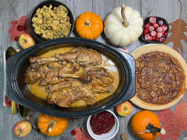 Food on a table shows turkey, cranberry, stuffing, and a pecan pie. Pumpkins and fall leaves surround them.