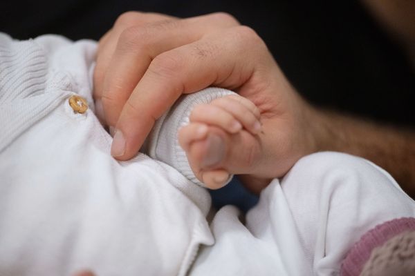 A white person holds the hand of a white baby. The baby is wearing white clothes. Their faces are not included in the image.