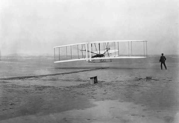 A plane is flying in the air lowly above the ground. A person stands next to the plane.