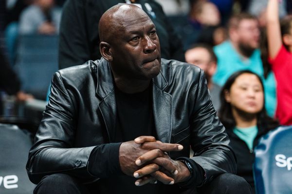 A Black man wearing a black leather jacket and pants is sitting down with his hands crossed in his lap. You can see multiple people sitting behind him in stands.