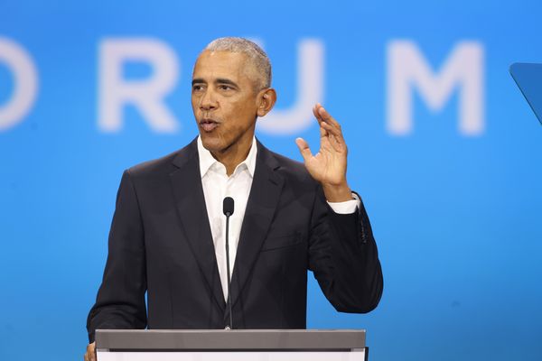 A Black man wearing a black suit raises his right hand as he speaks before a podium.