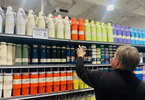 A white man stands before water tumblers in a range of different colors. A sign limits the amount that patrons can buy at the store.