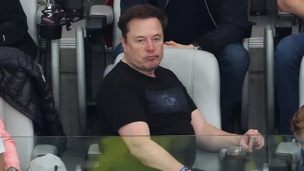 A white man wearing a black T-shirt stares foward. He is sitting amongst other people outside.