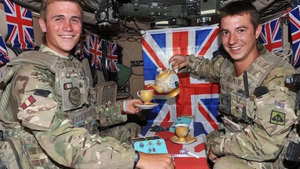 Two white people wearing camouflage uniforms are seated in front of British flags. The one on the left is holding a teacup on a saucer, while the one on the right is pouring tea from a teapot. 