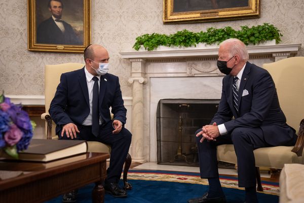 Two white men wearing masks and blue suits sit in white chairs.