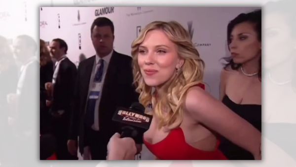 A white woman in a red dress smiles while talking into a microphone. A woman in a black dress stands slightly behind her. A person in a black suit stares at the camera. Other people stand on the other side of the person.