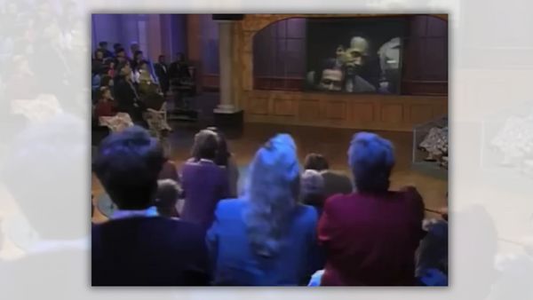 A crowd of people look at a TV screen that shows a Black man looking downcast.