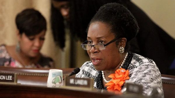 A Black woman speaks into a mic while sitting at a desk.