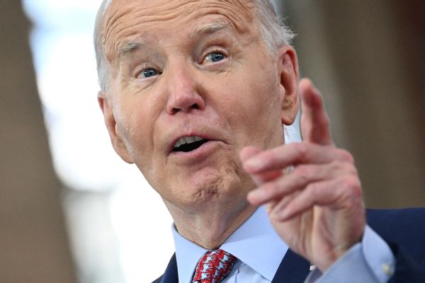 A white man wearing a blue suit points the index finger of his right hand in front of his face.