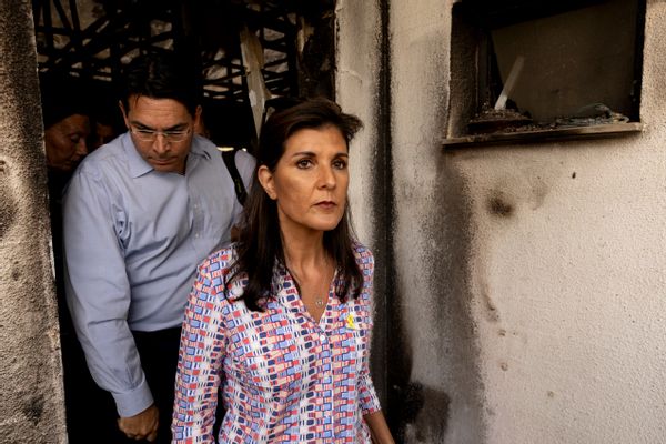 An Indian woman walks through a damaged house. A person walks behind her, and you can see other people behind them.